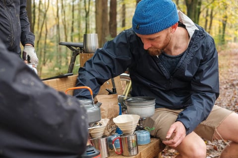 Man in a blue beanie makes filter coffee in woodland, with a camp stove, kettle and paper filters on a wooden bench.