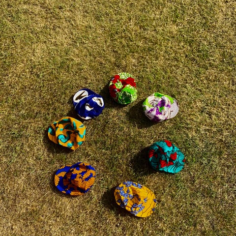 Seven Africap cycling caps in colourful Ankara fabrics laid out in a circle on dry grass.