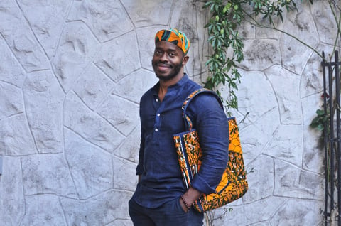A smiling man in an Africap Ankara-print cap carries a matching orange patterned tote bag against a stone wall.