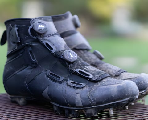 Pair of muddy Lake MX145 winter boots with dual BOA dials, seen from the front on a metal grid.