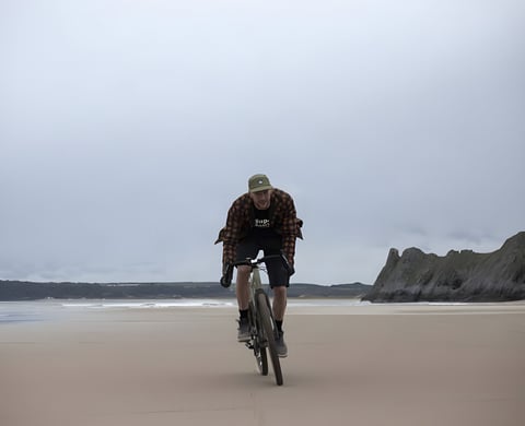 Rider in The Overland checked shirt and cap crosses a wide, empty beach on a gravel bike, with a rocky headland behind.