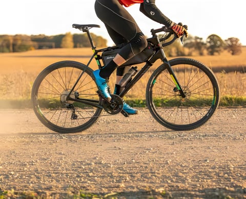 Rider on a Salsa Warbird gravel bike rolls along a dry gravel road past fields in low autumn light.