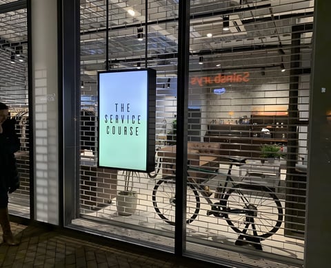 The Service Course Wilmslow shopfront after dark, with an illuminated sign and a bike visible behind the shutter.