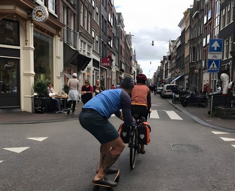 A skateboarder hangs on to a bike with orange panniers along a busy Dutch city street, an everyday adventure.