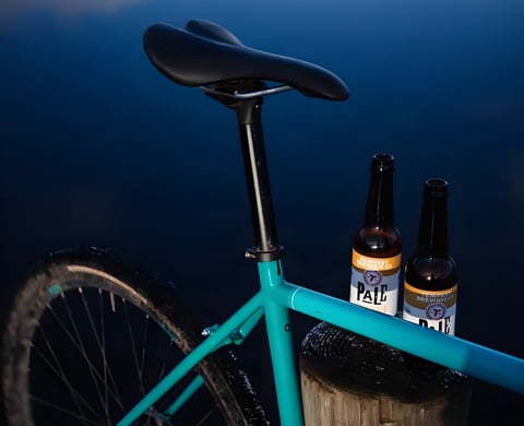 Two bottles of pale ale resting on the top tube of a teal gravel bike, a Muck n Mac Fest bikes-and-beer scene.