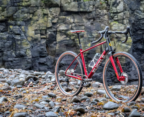 Red FUSTLE Causeway GR1 gravel bike with tan-wall tyres standing on a pebble shore strewn with seaweed.