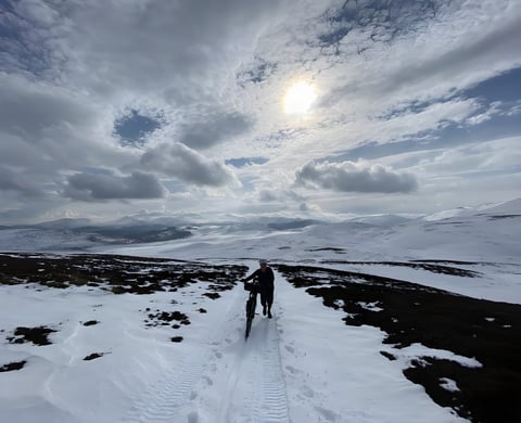 Lone rider pushes a loaded bike along a snowy track under a hazy sun, high on the Cairngorms bikepacking route.