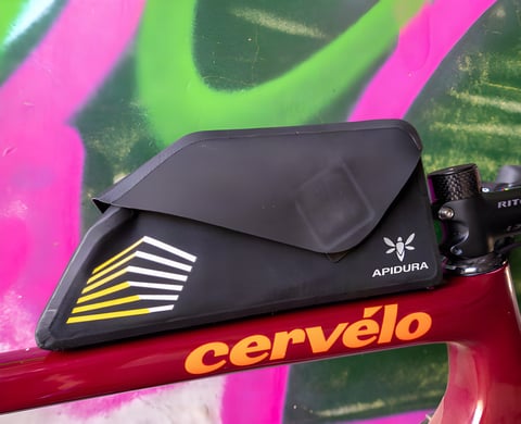 Apidura Racing bolt-on top tube bag with its flip-top cover on a maroon Cervélo frame against a graffiti wall.