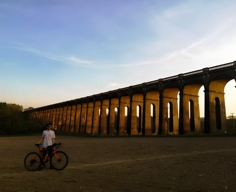 Ben Phillips stands with his red gravel bike in front of a long brick viaduct at dusk on a lockdown ride.