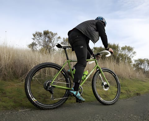Rider stands on the pedals of a green Ritchey Outback V2 steel gravel bike on a paved lane beside tall dry grass.
