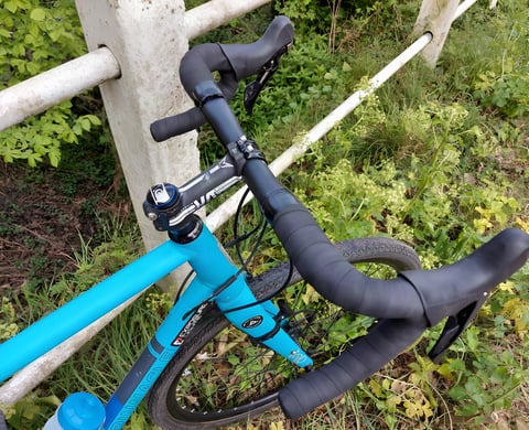 Genetic Driser 4-10 drop-riser bars with black tape on a blue Pinnacle gravel bike leant against a white rail fence.