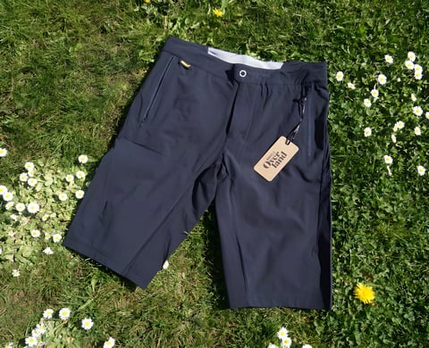 Overland Selector shorts in dark grey laid on grass among daisies, with the cardboard Morvélo Overland tag attached.
