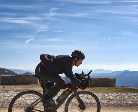 Garmin Edge Plus launch image: a rider on a gravel bike crosses a stony mountain road beside a low wall under blue sky.