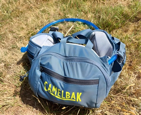Camelbak Repack LR 4 hydration waistpack in blue lying on dry grass, with the hose looped over the top.