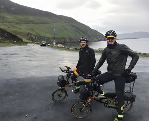 Two TransAtlantic Way riders on loaded Brompton folding bikes beside a green hillside and sea inlet on Ireland's west coast.