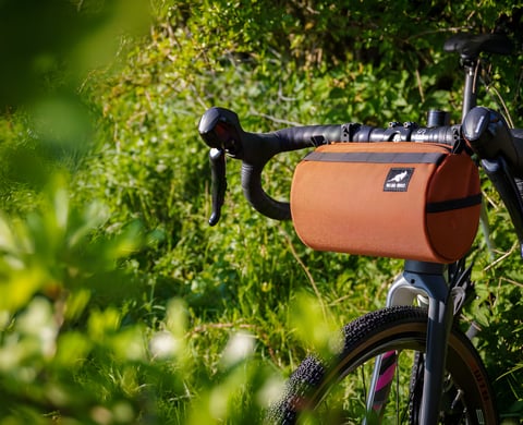 Wizard Works Lil Presto! barrel bag in rust strapped to the drop bars of a gravel bike, with green hedgerow behind.