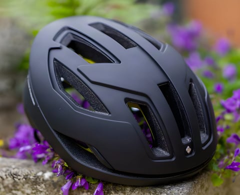 Sweet Protection Falconer II MIPS road helmet in matte black resting on a stone ledge among purple flowers.