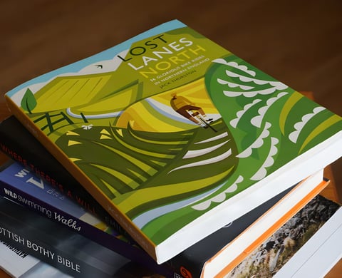 Lost Lanes North by Jack Thurston, an illustrated cycling guide, lying on a stack of books.