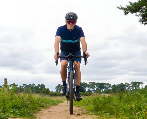 Rider wearing the Chapeau! Café Colour Block Jersey in Deep Ocean blue on a gravel bike, on a sandy track.