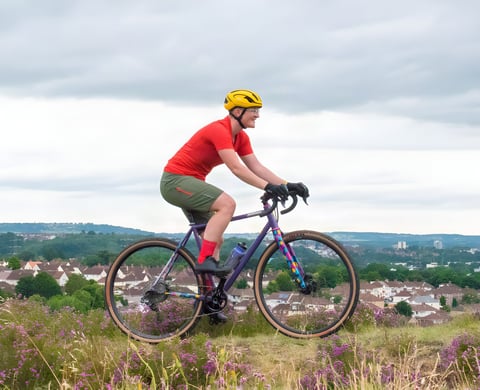 Katherine Moore rides a purple gravel bike through heather in Velocio TRAIL kit, red tee and khaki shorts, above a town.