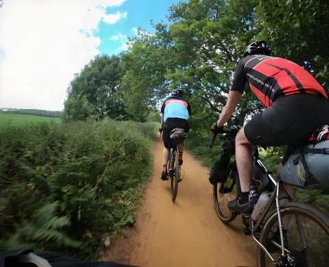 Two riders on loaded gravel bikes follow a sandy track between hedgerows and trees on a Norfolk rail trail.