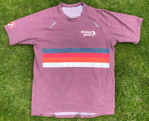 Stolen Goat Merino Gravel Jersey in washed-out red with retro stripes across the chest, laid flat on grass.