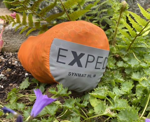 Exped SynMat HL M sleeping mat in its orange stuff sack, resting among ferns and plants.