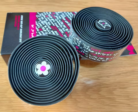 Two rolls of Kinesis Jo Burt signature bar tape in black with white script and a flower bar plug, on their box.