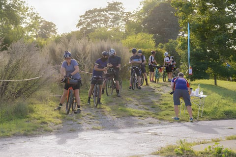 Great British Escapade riders wait behind the start tape on a grassy track in low evening sun.