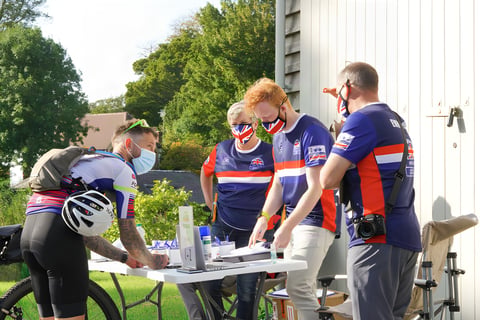 A rider signs on at a table at the Great British Escapade start as masked marshals in Union flag kit look on.