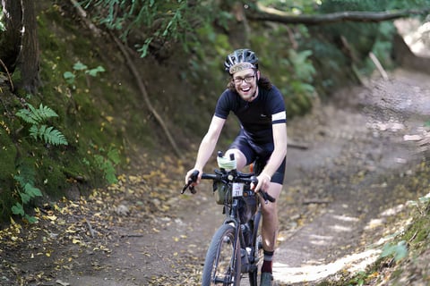 Smiling Great British Escapade rider climbs a sunken wooded track with a race number on the handlebar bag.