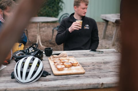 Tray of Portuguese custard tarts on a wooden table beside cycling helmets and a coffee at the Canterbury café stop.