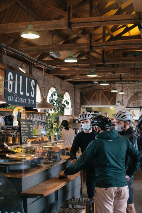 Riders in helmets and masks queue at the counter of Gill’s Café and Delicatessen in The Goods Shed, Canterbury.