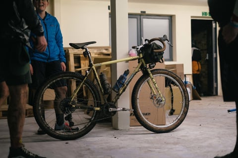 Mud-caked cream gravel bike with a bar bag propped by a pillar in the Brother Cycles warehouse in Faversham after the ride.