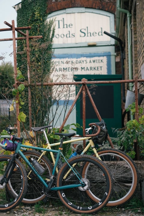 Two gravel bikes, one teal and one cream, leaning against railings outside The Goods Shed in Canterbury.