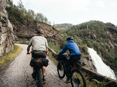 Two bikepackers ride a narrow cliffside road past a waterfall on the Lost Captures tour.