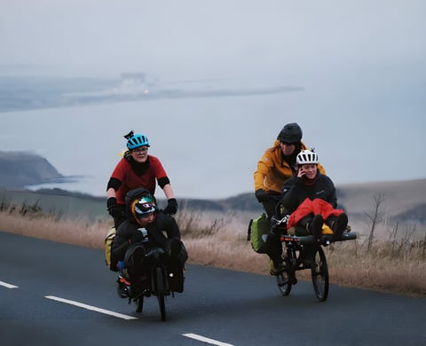 Four Adventure Syndicate riders share two cargo bikes on a coastal road above a grey sea during the Resolution Race.