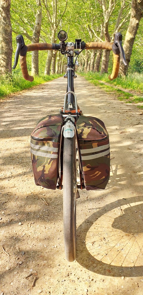 Front view of a drop-bar bike with camouflage Tim Tas &amp; Rek panniers on a shaded gravel track lined with trees.
