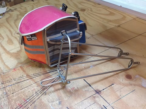 Tim Tas &amp; Rek handlebar bag in pink and orange beside a slim steel front rack on a workshop floor.