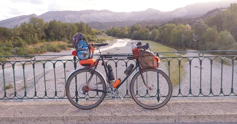 Touring bike with orange Tim Tas &amp; Rek bags and a camo bar bag leaning on a bridge railing above a river and mountains.