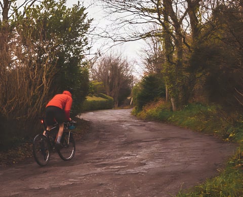Rider on a gravel bike climbs a potholed lane lined with bare trees, recceing a basket Everesting hill on the Wirral.