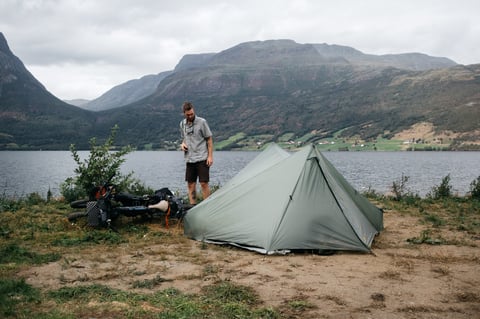 Green tent pitched on a lakeshore with a bike lying nearby and a rider standing below steep hillsides.