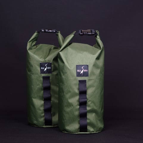 Two olive green Black Heron Fabrix roll-top dry bags with black webbing daisy chains against a dark background.