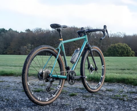 Stayer Groadinger UG steel gravel bike in teal with mud-caked tyres, parked on a gravel track beside a field.