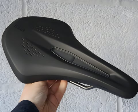 Fizik Terra Argo X3 gravel saddle held against a white wall, showing the short nose, cut-out insert and black cover.