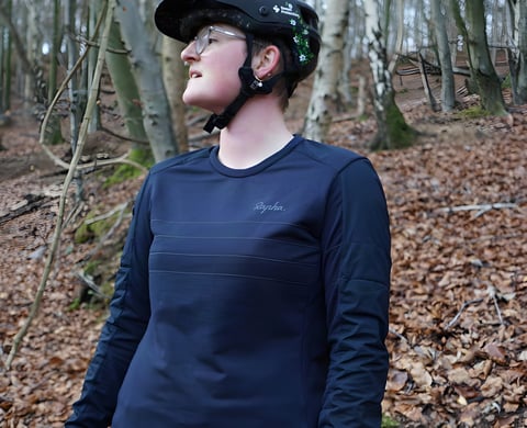 Rider in a navy Rapha Explore Long Sleeve Pullover and MTB helmet standing in autumn woodland.