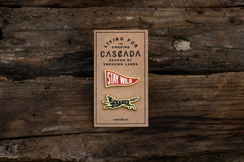Cascada enamel pin set on a card reading Stay Wild, a red pennant and a running fox, on weathered wood.
