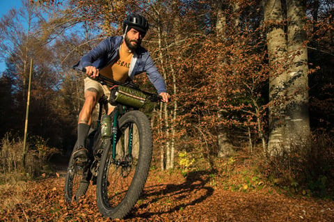 A rider in a Cascada top on a fat-tyre bikepacking bike with a green handlebar bag rides through autumn woodland.