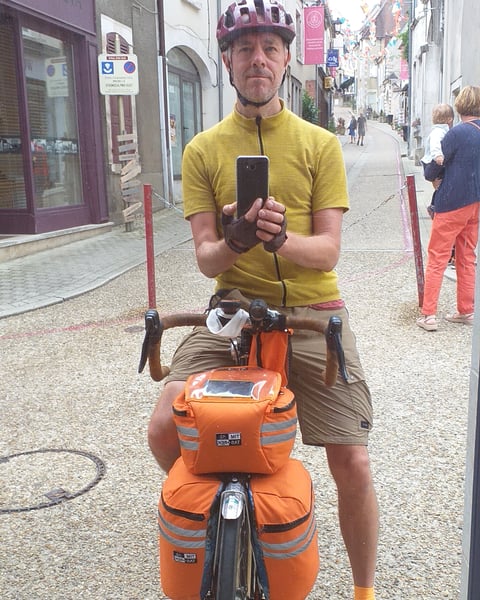 Cyclist in a yellow jersey takes a photo astride a touring bike loaded with orange Tim Tas &amp; Rek bags on a town street.