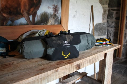 Apidura Backcountry handlebar pack and accessory pocket resting on a wooden table in a stone building.